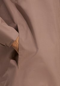 Tissu lisse de couleur marron clair avec une poche sur le côté gauche. La couture est visible, avec un éclat subtil et une texture douce.