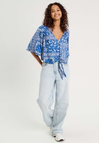 Haut bleu à manches courtes arborant des motifs paisley et floraux blancs, avec un col en V et un détail noué à la taille. Pantalon en jean délavé porté.