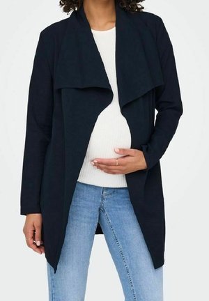 Schwangere Person in hellblauen Jeans, weißem Oberteil und offenem dunkelblauem langem Cardigan, die Hand auf dem Bauch ruhend.