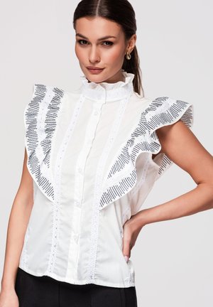 Femme aux cheveux foncés porte un chemisier blanc sans manches avec un bord festonné à motif et un col montant, posant la main sur la hanche devant un fond uni.