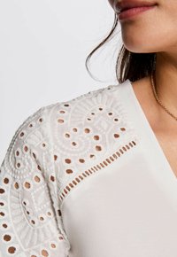 Weiße Bluse mit bestickten Spitzenärmeln und Ausschnittmustern, V-Ausschnitt und glatter Textur am Oberteil. Feine Details sichtbar.
