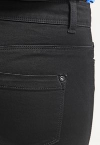 Jeans de mezclilla negra con un diseño clásico, bolsillo trasero con un detalle de botón metálico y costuras en contraste.