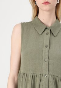 Robe sans manches vert olive avec un col, des boutons sur le devant, et une taille élastiquée. Le tissu présente une texture subtile.