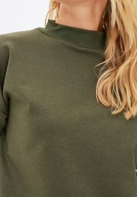 Olivgrön luvtröja med hög hals. Mjukt tyg, figursydd design, inga synliga mönster eller hårddetaljer.