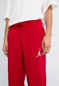Piros, pamutból készült melegítőnadrág, elasztikus derékpánttal és húzózsinórral, valamint egy fehér Jumpman logós díszítéssel a combján.