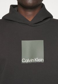 Donkere hoodie met een groen vierkant en witte "Calvin Klein"-logo op de borst, gedragen door een persoon met een donkere huid.