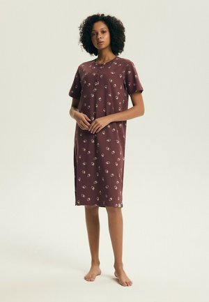 Kobieta stojąca boso, ubrana w luźną, sięgającą kolan bordową sukienkę z krótkimi rękawami i małymi jasnymi kwiatowymi wzorami.