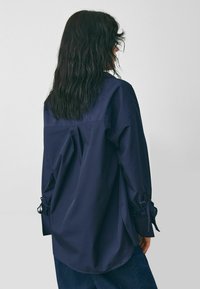 Námornícka modrá oversized košeľa vyrobená z hladkej tkaniny, s chrbátom yoke, záhybmi a nastaviteľnými viazaniami na rukávoch pre zvýraznenie detailu.