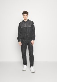Svart träningsset bestående av en jacka med dragkedja som har ett mönstrat övre parti, elastiska manschetter och matchande byxor. Vita sneakers fulländar looken.