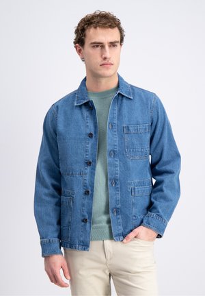 Mężczyzna w niebieskiej kurtce dżinsowej na zielonym swetrze i beżowych spodniach, stojący z jedną ręką w kieszeni na jednolitym tle.