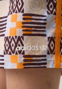 Pantalones cortos con estampado sobre base rosa, con formas geométricas en naranja, azul y marrón; incluye logo de Adidas bordado en blanco y detalles en naranja.