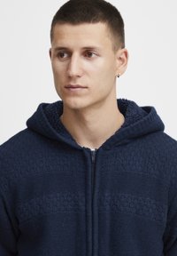 Tamsiai mėlynas zip-up džemperis su gobtuvu, pagamintas iš tekstūruoto audinio. Yra pamuštas gobtuvas ir horizontalaus rašto akcentai ant kūno.