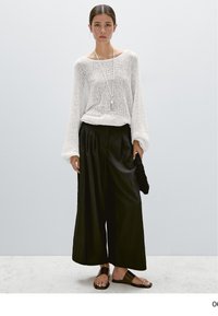 Frau trägt weißen locker gestrickten Pullover, schwarze weit geschnittene Hose, schwarze Sandalen, hält schwarze, strukturierte Clutch vor schlichtem hellem Hintergrund.