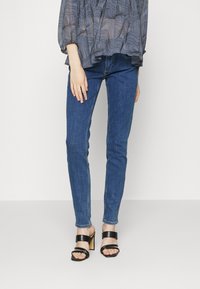 Blue denim skinny jeans with a smooth texture; paired with black heeled sandals featuring two wide straps, set against a neutral background.