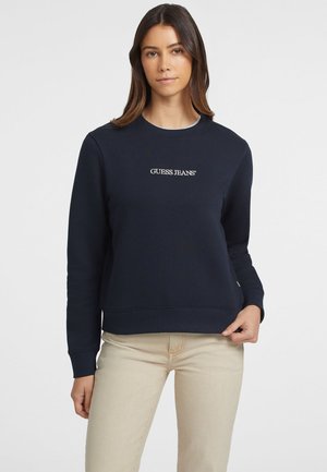Navy sweatshirt with a round neck, ribbed cuffs, and a subtle "GUESS JEANS" logo embroidered on the front, paired with beige pants.