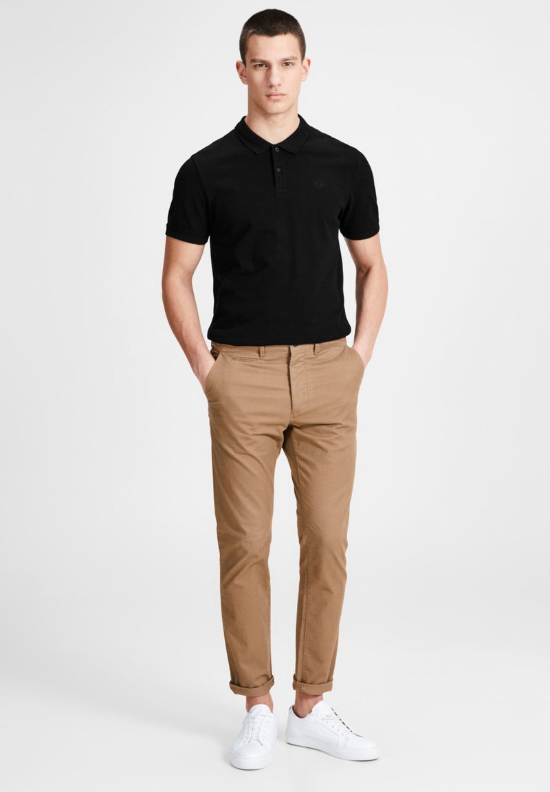 Schwarzes Kurzarm-Polohemd mit Kragen, kombiniert mit beigefarbenen Chinos und weißen Sneakers. Lässige Passform mit hochgekrempelten Säumen an der Hose.