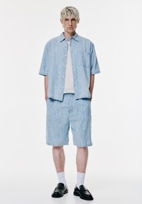 Camicetta a righe azzurro chiaro con maniche corte, pantaloni corti abbinati, maglione bianco sotto, calze bianche e mocassini neri; stile casual, vestibilità rilassata.