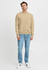 Beige sweatshirt med rund halsudskæring, struktureret stof og cirkulært logo-detail, parret med lyseblå jeans og hvide sneakers med sorte striber.