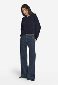 Marineblauer Oversized-Pullover mit rundem Halsausschnitt, kombiniert mit dunkelblauen weit geschnittenen Jeans. Die Accessoires bestehen aus einem braunen Gürtel und schwarzen Schuhen.