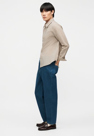 Νεαρό άτομο με μαύρα μαλλιά φορώντας μπεζ πουκάμισο με κουμπιά, μπλε φαρδιά τζιν, λευκές κάλτσες και σκουρό καφέ μοκασίνια, στέκεται πλευρικά.