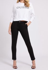 Felpa bianca corta con "GUESS" in testo argentato; abbinata a pantaloni neri slim-fit; il modello indossa tacchi neri con cinturini; texture del tessuto liscia.