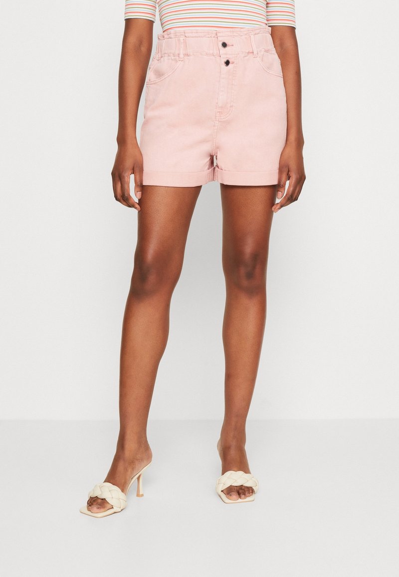 Femme portant un short en denim rose clair à taille haute, un haut rayé, et des sandales à talons hauts en tissage crème, se tenant devant un fond uni.