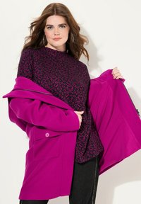 Fuchsiový kabát s velkými kapsami a zapínáním na knoflík, navrstvený na černém topu s růžovým leopardím vzorem, spárovaný s tmavými džíny.