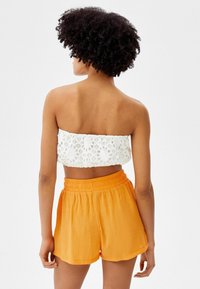 Mujer con cabello rizado que lleva una blusa bandeau de encaje blanco y pantalones cortos de talle alto de color naranja brillante, de pie de espaldas contra un fondo liso.