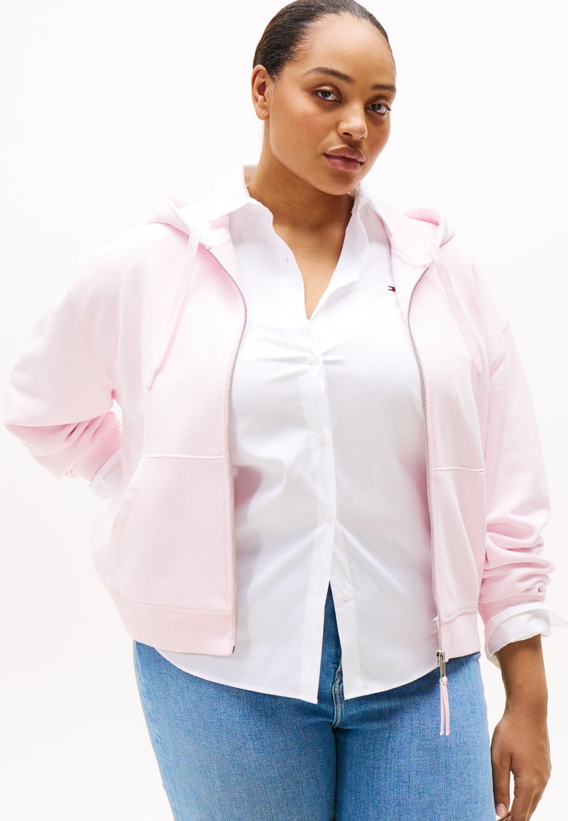 Lichtroze hoodie met rits en zakken, gecombineerd met een witte overhemd. Voorzien van een capuchon met trekkoord en een textuurstof. Blauw jeans maken de look compleet.