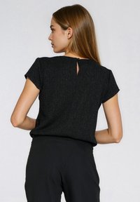 Czarny top z krótkim rękawem o fakturowanym wzorze, z wycięciem w kształcie łezki z tyłu zapinanym na guzik. W zestawie z czarnymi spodniami.