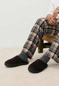 Hnedé kordové papuče s textúrovaným vrchom a čiernou gumovou podrážkou, nosené so šedými ponožkami a nohavicami s kockovaným vzorom.