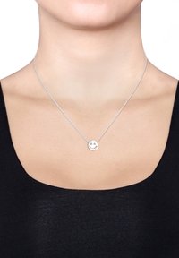 Sølvring med en rund medaljon med et smilende ansigt design. Kæden er delikat og fint sammenkoblet, båret over en sort top.