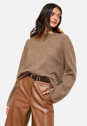 Brauner gestrickter Pullover mit übergroßen Ärmeln, kombiniert mit hochtaillierten braunen Kunstledershorts und einem dunklen Gürtel. Glatte Textur und lässige Passform.