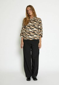 Blusa stampata con motivi ondulati beige, marrone e neri, colletto e maniche a tre quarti, abbinata a pantaloni larghi neri e scarpe.