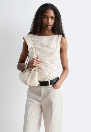 Joven mujer con cabello rizado que lleva una blusa sin mangas estampada y pantalones claros, sosteniendo un bolso beige con una mano en el bolsillo.