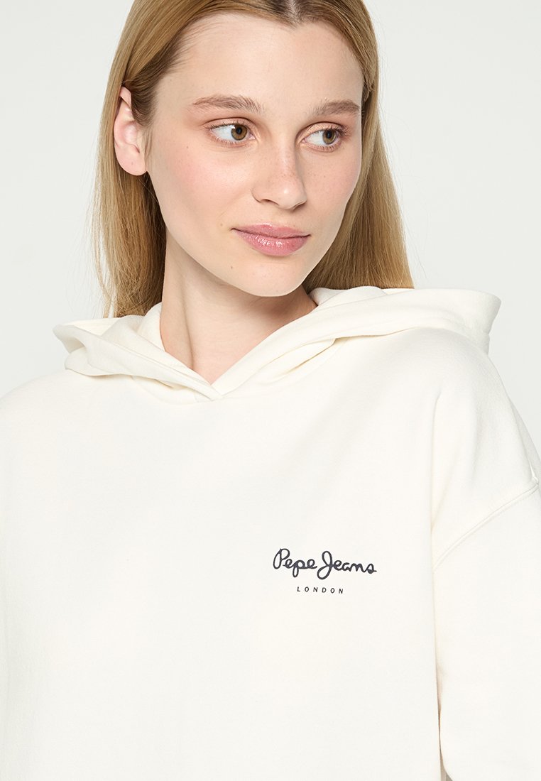 Sudadera color crema hecha de una tela suave, con un bolsillo tipo canguro, capucha ajustable y un pequeño logo negro en el lado izquierdo del pecho.