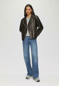 Schwarze Lederbikerjacke mit silbernen Reißverschlüssen, graues T-Shirt darunter, kombiniert mit hellblauen Schlagjeans und silbernen Sneakers.