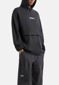 Persona che indossa un cappuccio nero con la scritta "INFINITY" e pantaloni neri, con le mani in tasca, rivolta in avanti su uno sfondo chiaro e semplice.