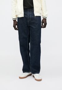 Mørkeblå denimjeans med avslappet passform, med glidel�ås foran og kontrastfargede sømmer. Brukes med en lys kremfarget jakke og hvite joggesko.