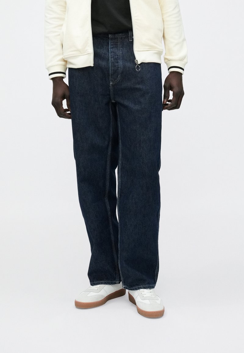 Mørkeblå denimjeans med avslappet passform, med glidelås foran og kontrastfargede sømmer. Brukes med en lys kremfarget jakke og hvite joggesko.