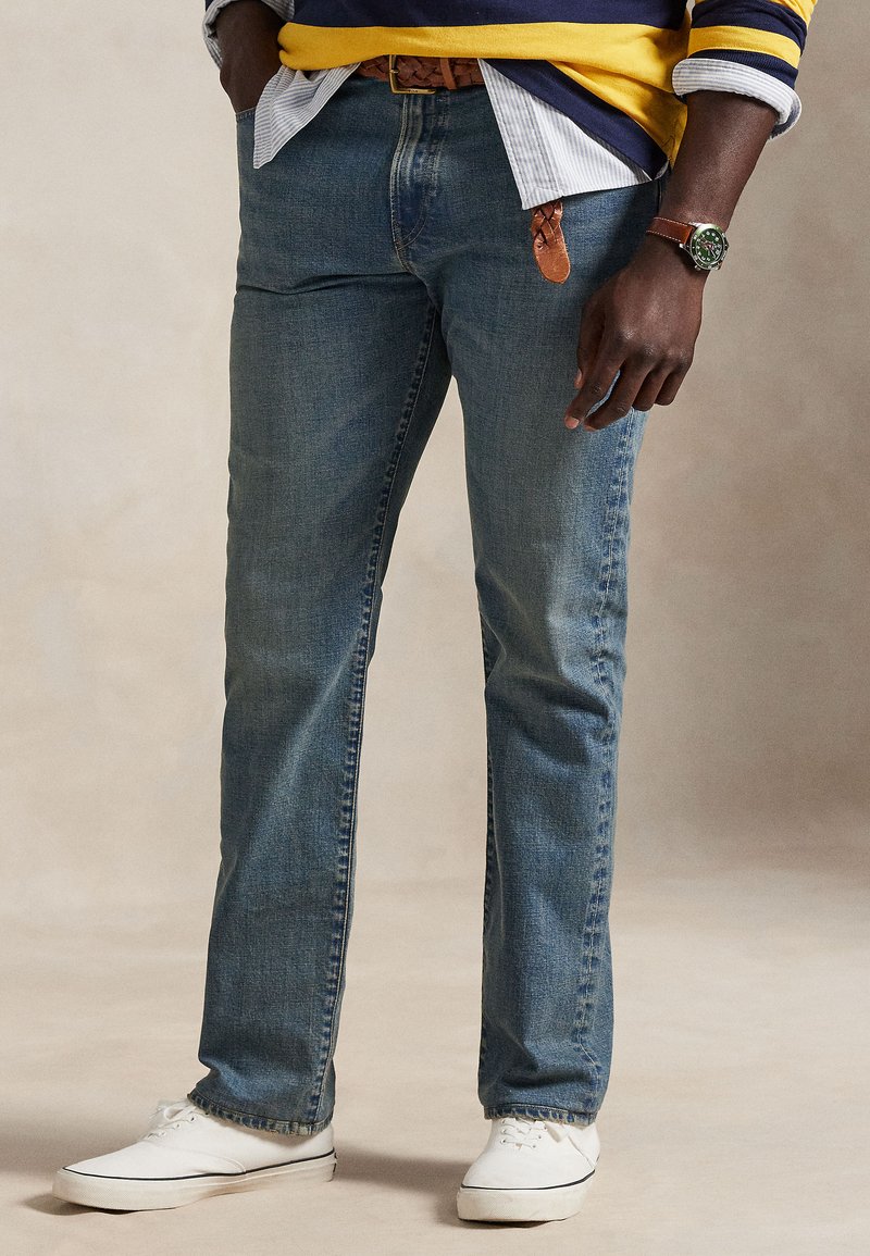 Person iført blå jeans, hvide sneakers, en stribet skjorte og en gul og navy sweater med et brunt læderbælte og et armbåndsur.