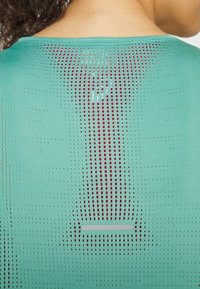 Zielony top sportowy wykonany z teksturowanej, oddychającej tkaniny z perforacjami. Tył zawiera czerwone akcenty i wzór w poziome paski.