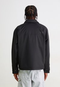 Veste noire imperméable avec un col, des manches longues et des boutons-pression. Présente un logo visible sur le haut du dos. Texture lisse.