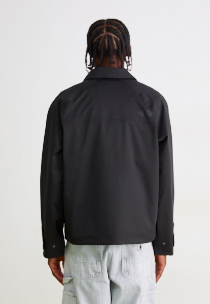 Schwarze wasserdichte Jacke mit Kragen, langen Ärmeln und Druckknöpfen. Verfügt über ein sichtbares Logo auf dem oberen Rücken. Glatte Textur.