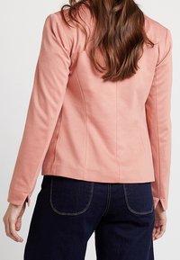 Vaaleanpunainen tiukka takki, jossa on pitkät hihat, teksturoitu kangas ja takasaumat, yhdistettynä tummansinisiin farkkuihin, joissa on näkyvät ompeleet.