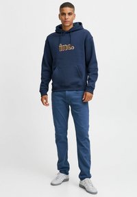 Felpa con cappuccio blu navy con tasca frontale, caratterizzata dal logo "inc." in testo colorato. Indossata con jeans blu e sneakers grigie.