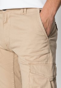 Close-up van een persoon die beige cargobroek draagt met een hand in de voorzak, waarbij de gedetailleerde stiksels en de textuur van de stof zichtbaar zijn.