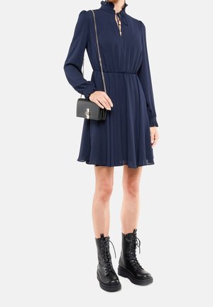 Femme portant une robe longue bleu marine à manches longues avec un col à volants, des bottes de combat noires à lacets et un petit sac bandoulière noir avec une anse chaîne.