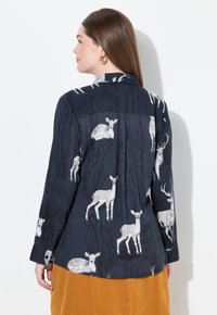 Donna con lunghi capelli castani che indossa una blusa blu navy con stampe di cervi bianchi e una gonna senape, rivolta a sinistra su uno sfondo bianco.