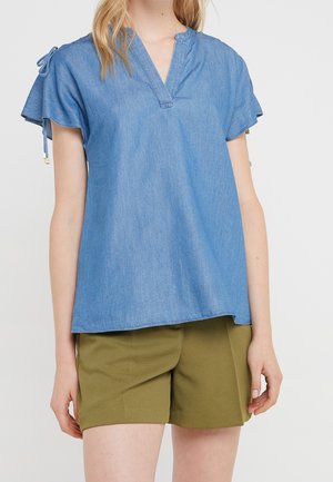 Femme portant un chemisier en denim bleu à manches courtes avec une encolure fendue et un short vert olive sur un fond uni.